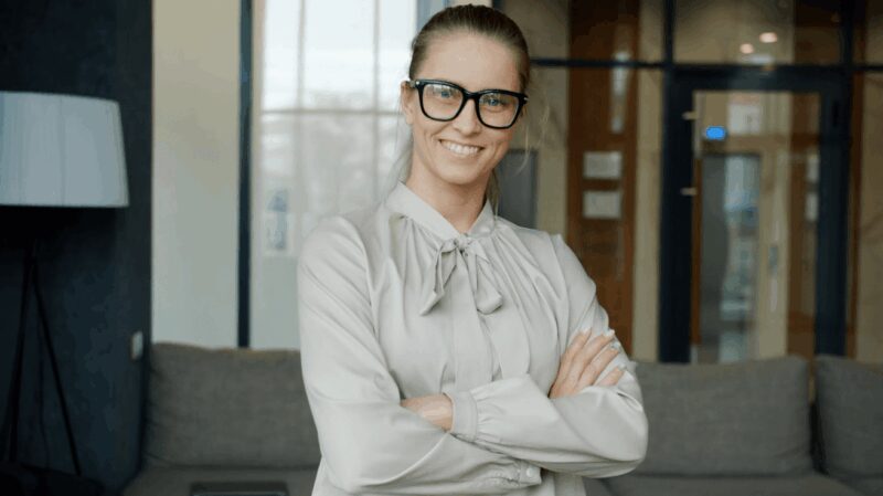 Žena u poslovno ležernom outfitu sa svetlom bluzom na vezivanje i naočarima, nasmejana i sa rukama prekrštenim ispred kancelarije.